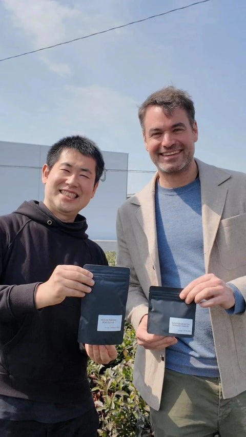 Two men standing outdoors in a tea plantation holding matcha pouches, with a large white warehouse building and blue sky in the background.