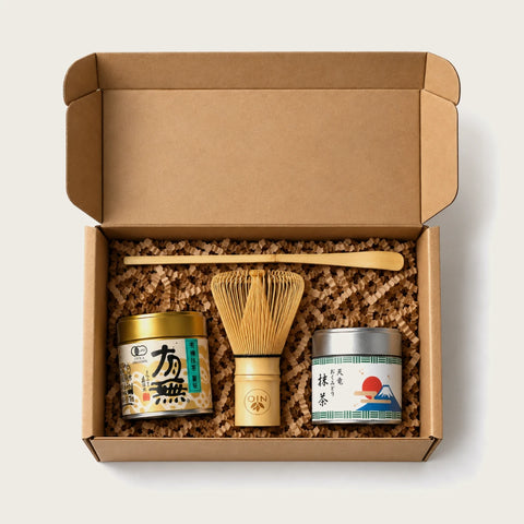 An open kraft cardboard gift box filled with brown crinkle paper, containing a bamboo whisk, a bamboo scoop, and two small tea tins with Japanese labels, photographed from above on a light neutral background.